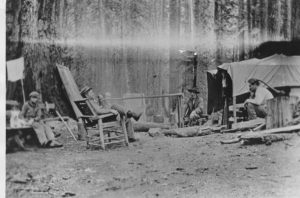 A man leaning back in a chair smoking a pipe with four other men sitting around a campsite.