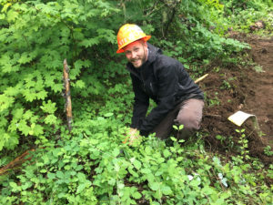 A man in rain jacket and hard hat kneeling in green foliage.