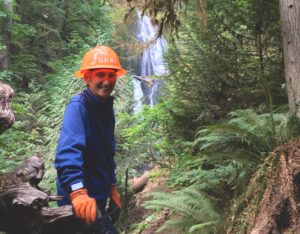 A woman in a hard hat on a narrow trail with a waterfall behind her. 