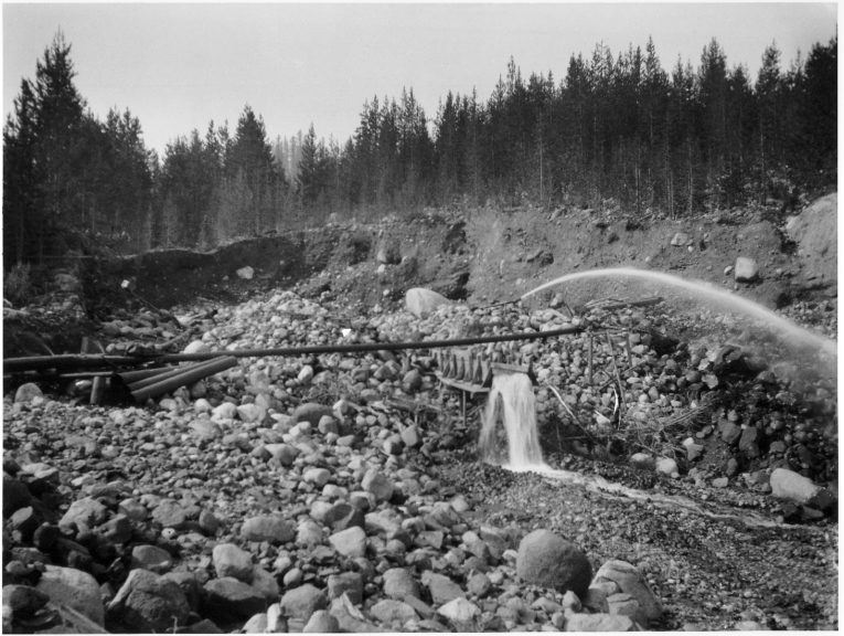 A black-and-white photo of a pipe spewing water and a sluice box or flue with water cascading off it in the midst of an acre of rocks with newly exposed cliffs in the background and coniferous trees rising above the cliffs.