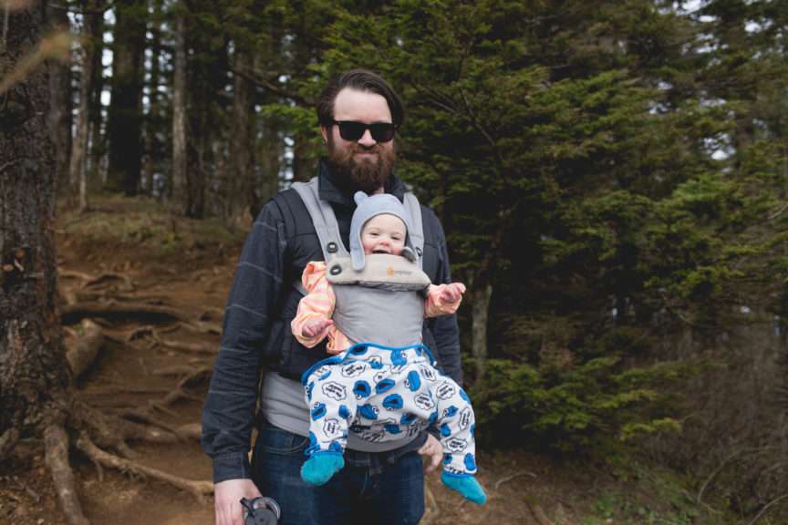 A man with a baby—who seems to be wearing voluminous pants—in a baby carrier on his chest, standing against a backdrop of a coniferous forest,
