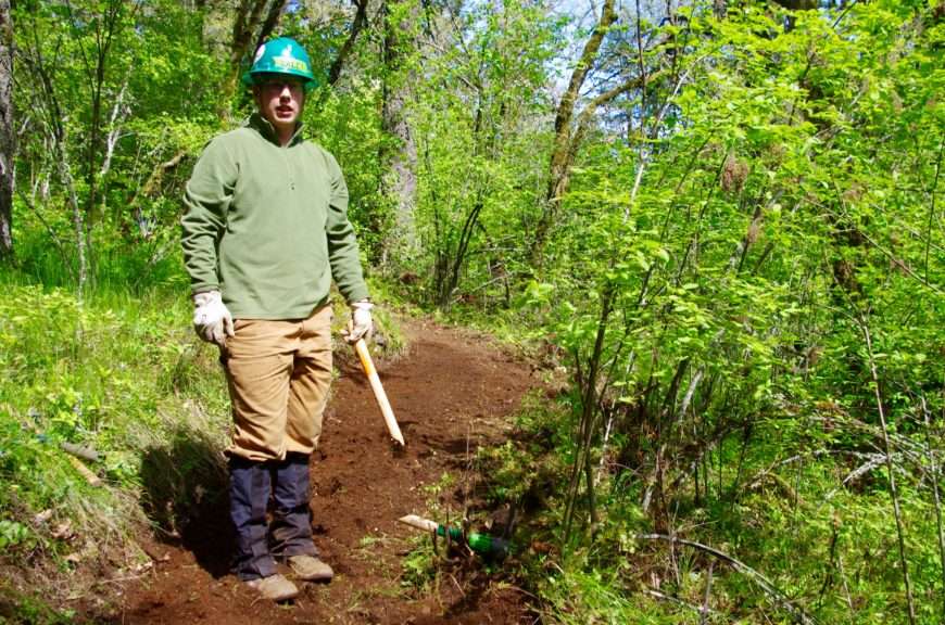A trail worker in a green hard hat and wearing gaiters stands in the middle of a dirt trail holding the broken=off handle of a trail tool. 