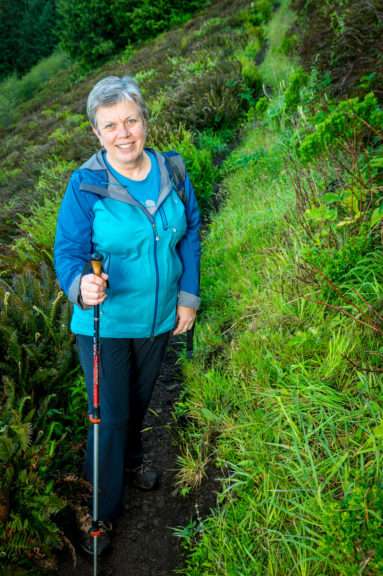 A woman stands on a narrow trail crossing a steep green slope, holding a hiking stick in one hand.