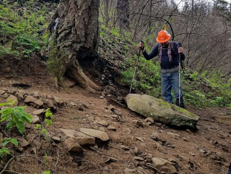 A trail worker in a hard hat stands looking down at a large rock.