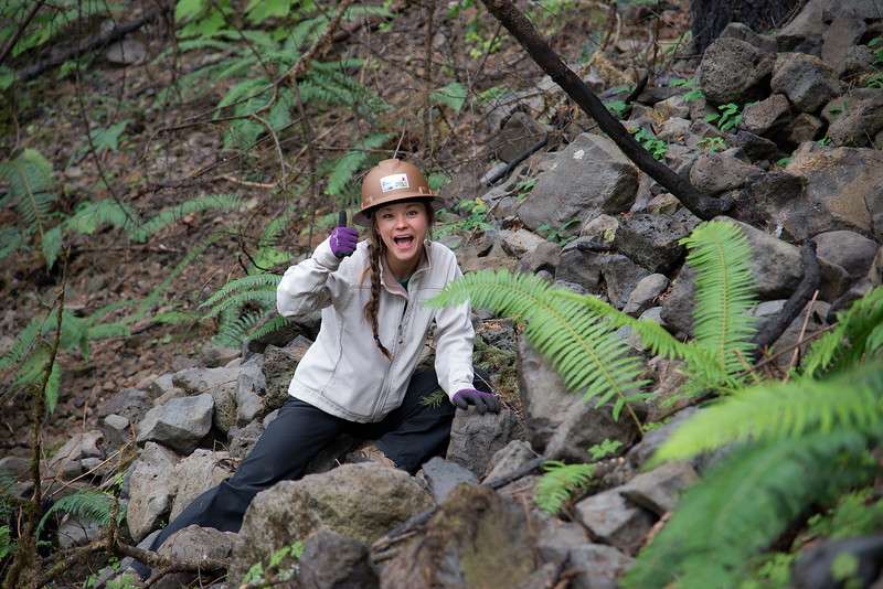 A hard-hatted woman on a rocky slope, giving a thumbs-up.