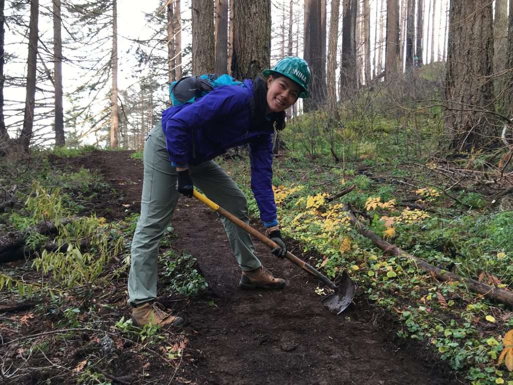 A woman in a hard hat leans over a trail in a wooded area while pulling the blade of a long-handled tool across the tread.