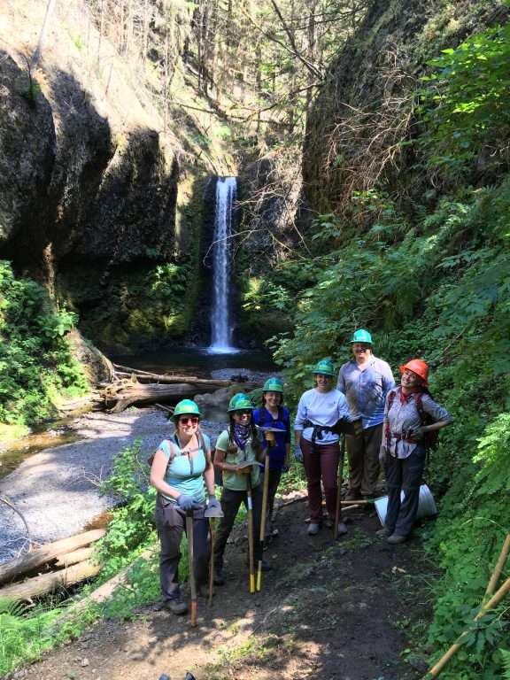 Six hard-hatted workers with tools in hand pose on a trail with a waterfall in the background. 