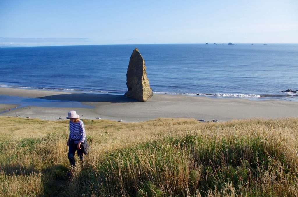 A woman walking across grassy dunes in the foreground, a tall sea stack casting a long shadow on a beach in the middle ground, and rocky islands in the ocean in the background.