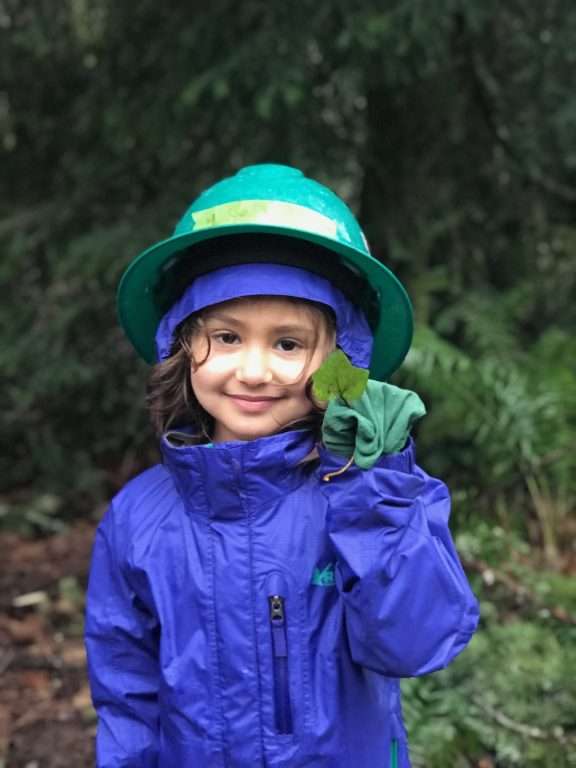 A young girl in a green hardhat holds up an ivy leaf.