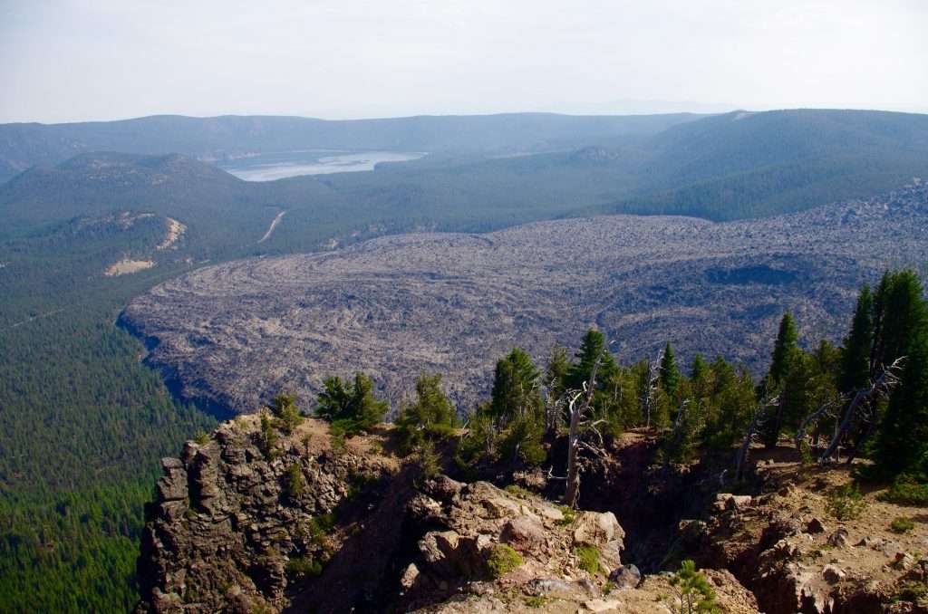 A cliff in the foreground, the grayish-black swirl of an immense lava flow far below, a lake beyond, and a caldera rim in the distance.