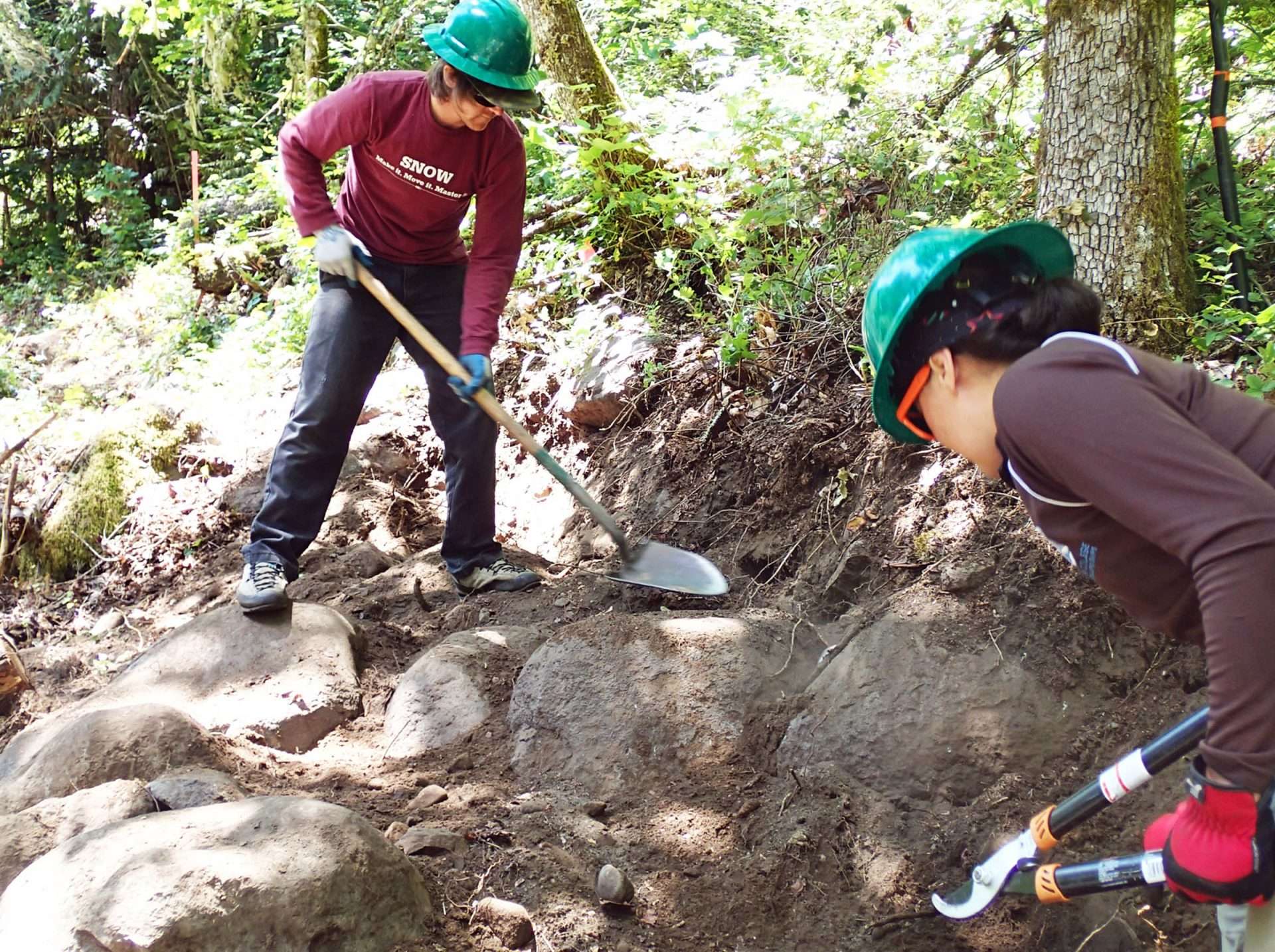 Two workers, one with a shovel and one with loppers, working on a section of trail that has more boulders than dirt.