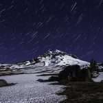 Star trails in a dark night sky appear above a snowy mountain peak.