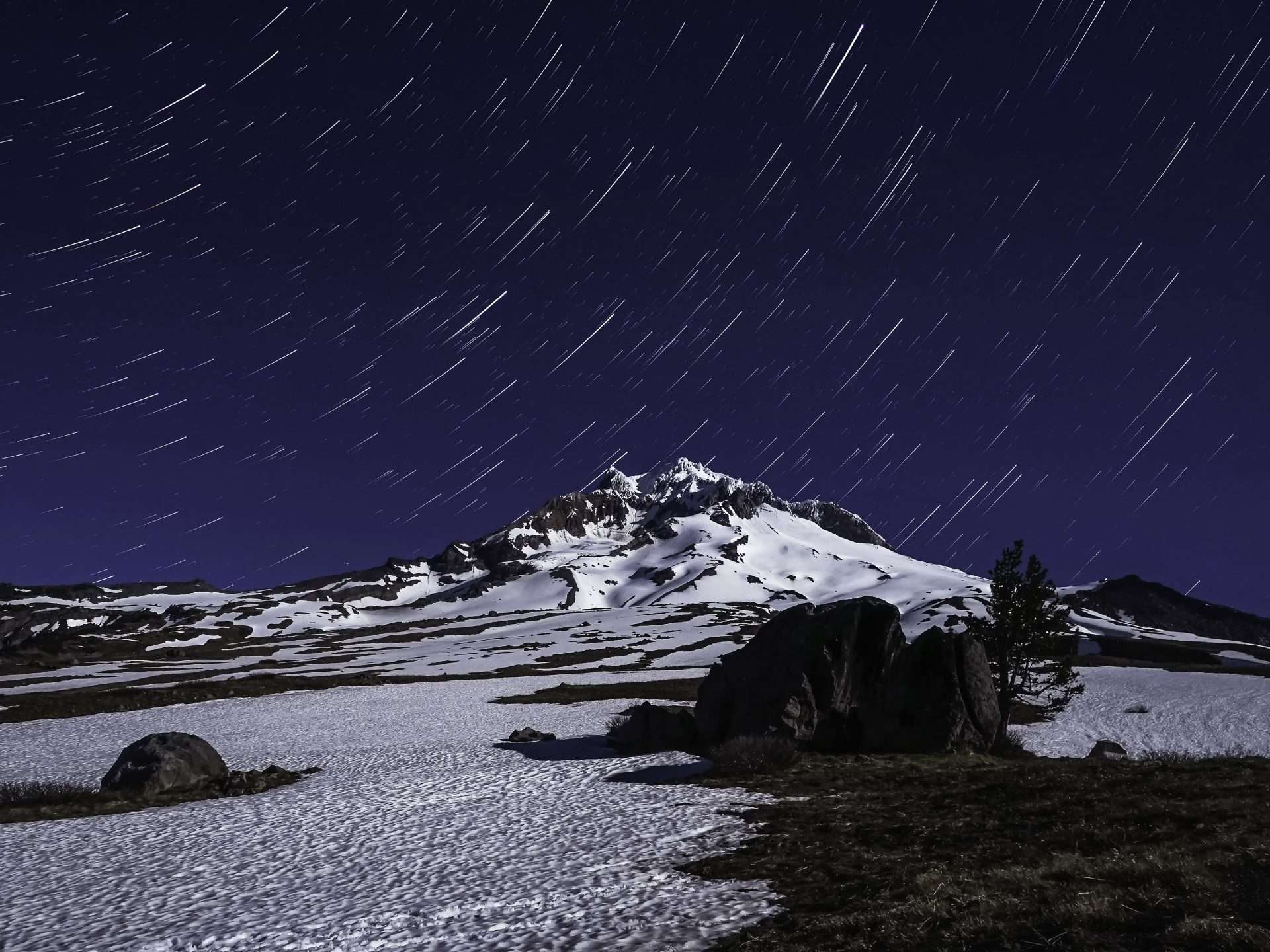 Star trails in a dark night sky appear above a snowy mountain peak.