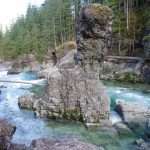 A rock pillar stands up from the middle of a greenish-blue river bordered by light grey rocks and dark green coniferous forest.