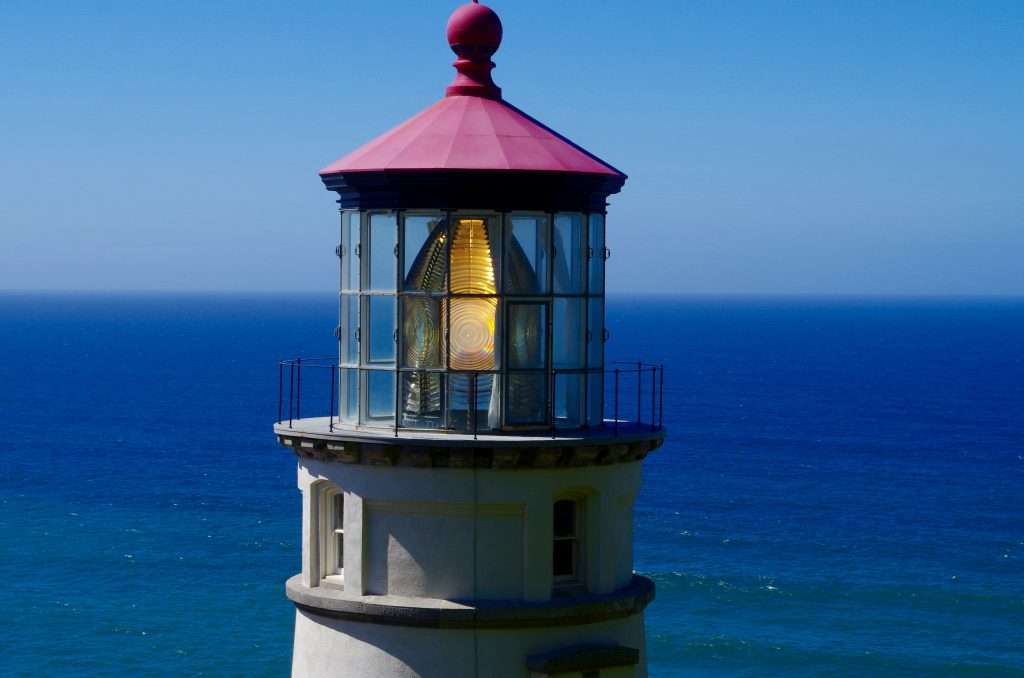A brightly lit lens under the red roof of a lighthouse with the ocean behind.