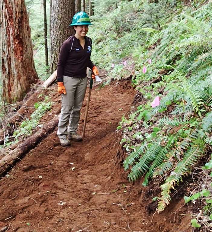A trail worker in a green hard hat stands on a newly restored section of trail on a forested hillside.