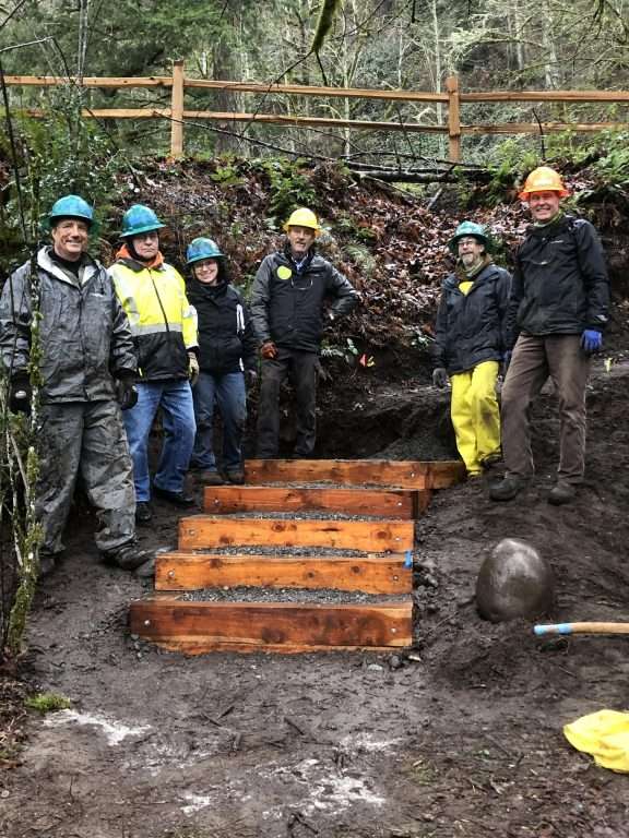 Six trail workers in hard hats and rain gear pose around a half-completed set of steps on a trail In a wooded area.