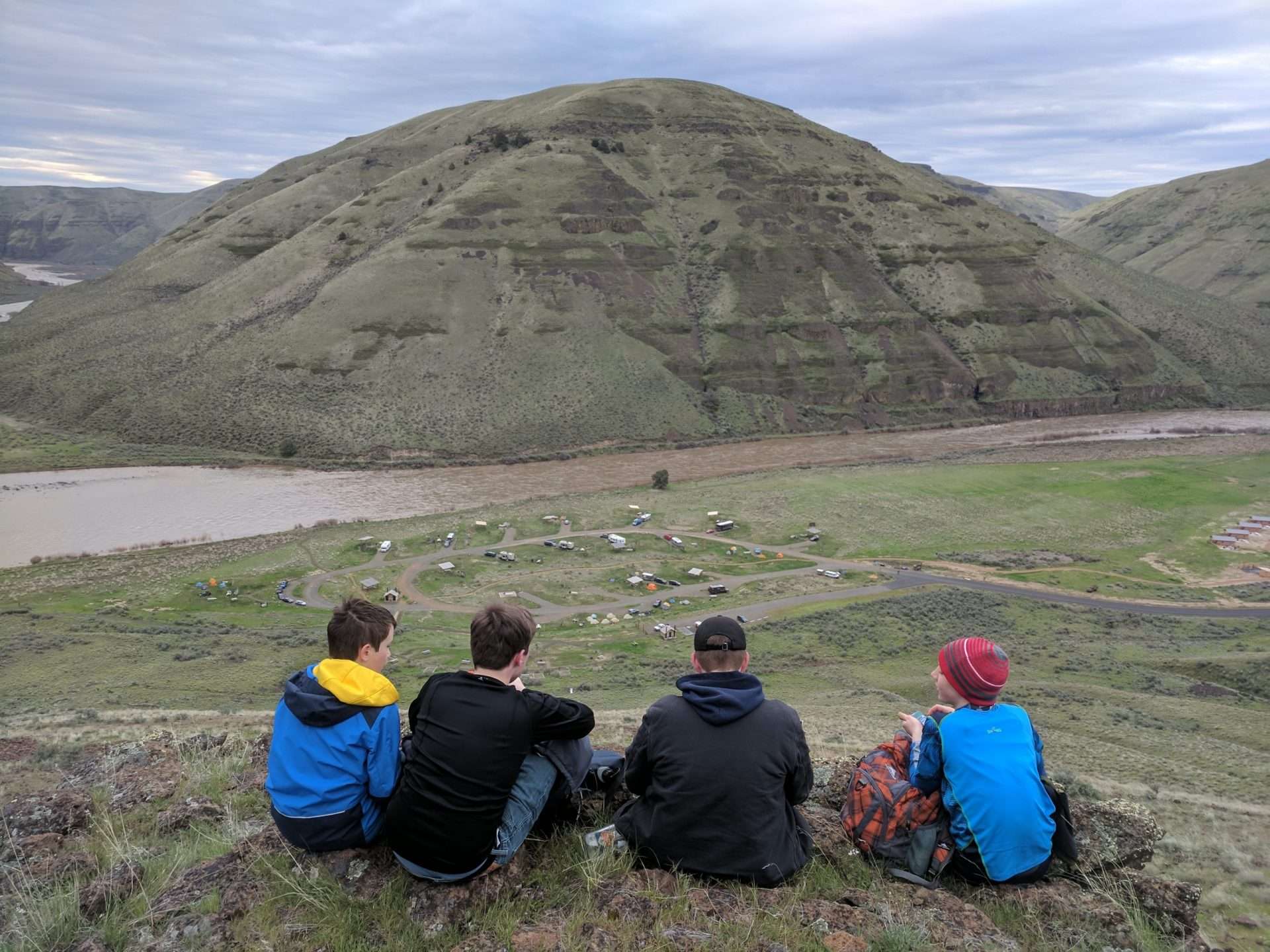 Four boys sitting on a hill overlooking a campground next to a muddy river, with a treeless slope across the river.