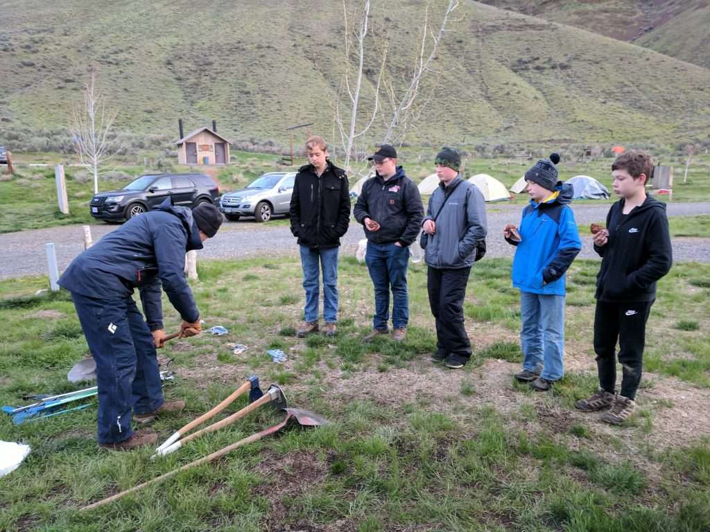 Five young boys stand in a line and listen to a man demonstrate how to use a shovel; another shovel and two hoes lie on the ground.