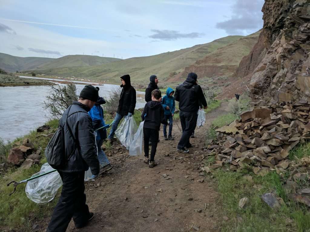 A group of boys and a couple of adult men carrying large plastic bags and trash pickers stand on a trail below a rocky cliff with a river running to the left.