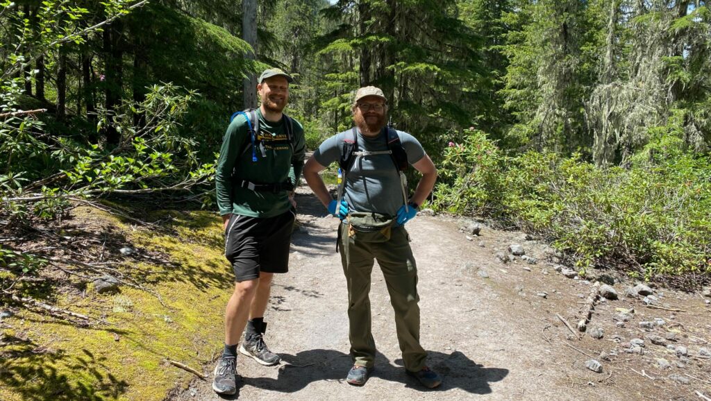 Two wilderness volunteers stand smiling next to each other on a sunny hiking trail
