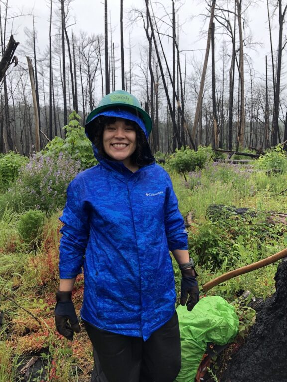 In front of a stand of burned trees and green under growth, a person in a blue rain jacket, black pants and a green hard hat smiles at the camera.